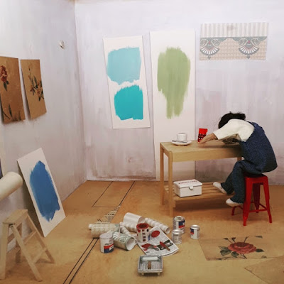 1/12 scale modern miniature scene of a room with paint and wallpaper swatches on the wall. In the middle of the room is a woman asleep at the workbench.