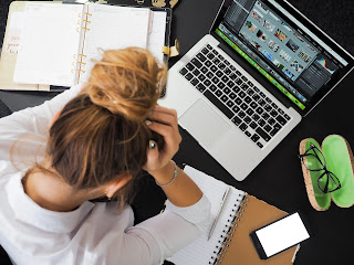 Woman holding head sitting in front of laptop with papers and books all around