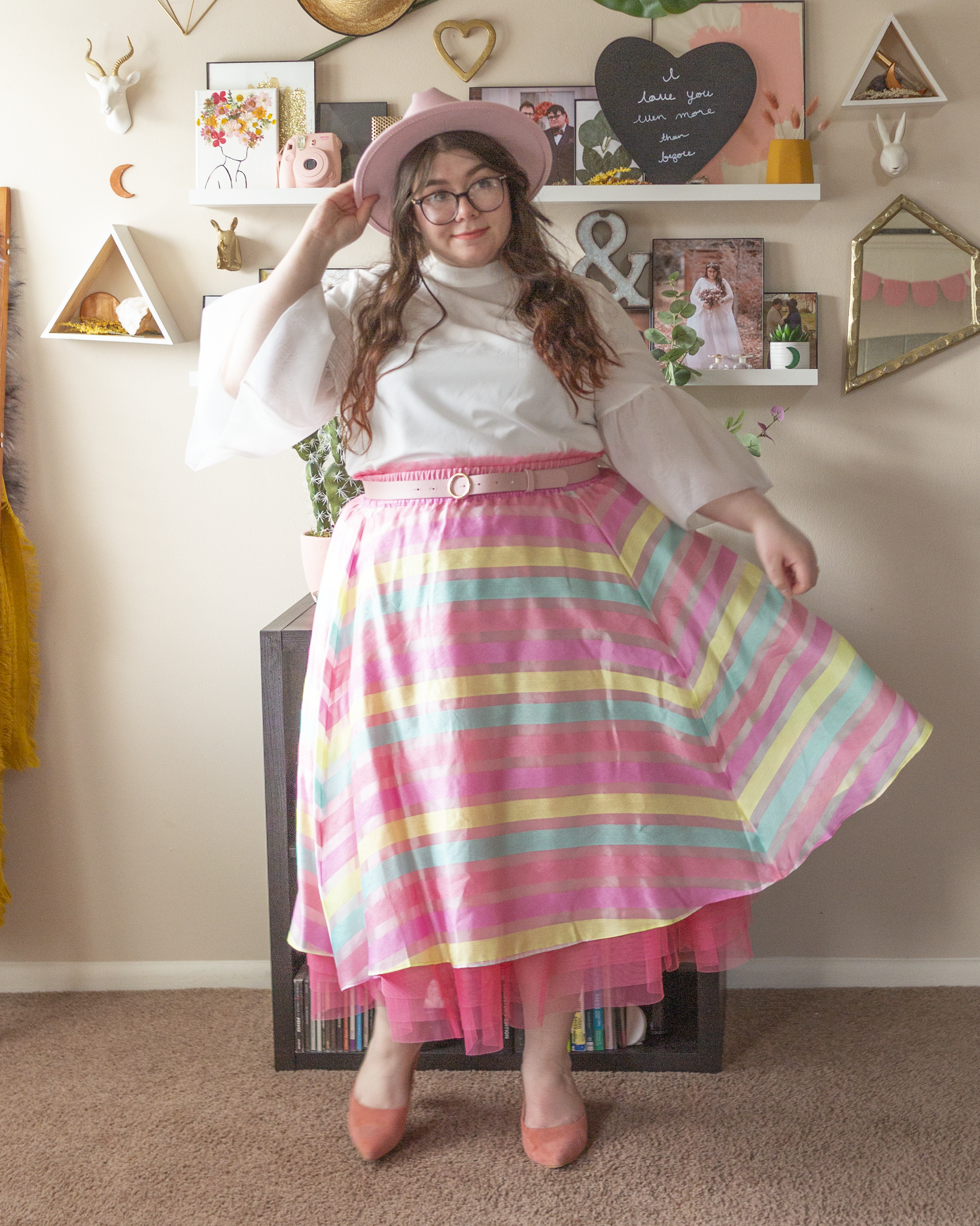 An outfit consisting of a wide brim pastel pink fedora, white short sleeve blouse with sheer organza bell sleeves tucked into a pastel pink, yellow and blue tulle and organza midi A-line skirt and pastel pink suede heeled clogs.