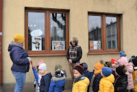 Grupa przedszkolaków z balonikami stoi przed okani biblioteki w Zelowie i ogląda wystawę.