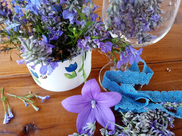 Composición de flores azules y lavanda en copa de cristal y taza, acompañado de cinta de puntilla azul para formar un bodegón.
