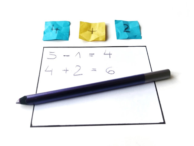 zabawy matematyczne dla przedszkolaków kartka z zapisanymi działaniami i kolorowa kredka leżą na pierwszym planie, za nią widac wylosowane karteczki z liczbami i znakiem dodawania
