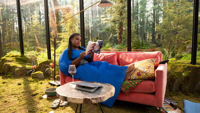 Personagem Sasha de SOLOS (Uzo Aduba) deitada num sofá lendo um livro dentro de uma casa de vidro cercada por uma floresta