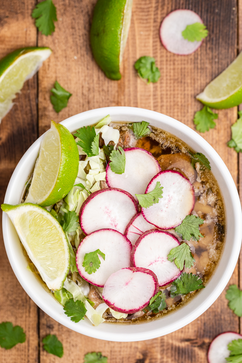Overhead view of Keto Pork Pozole Verde in a white bowl with sliced radish, cabbage, and lime wedges.