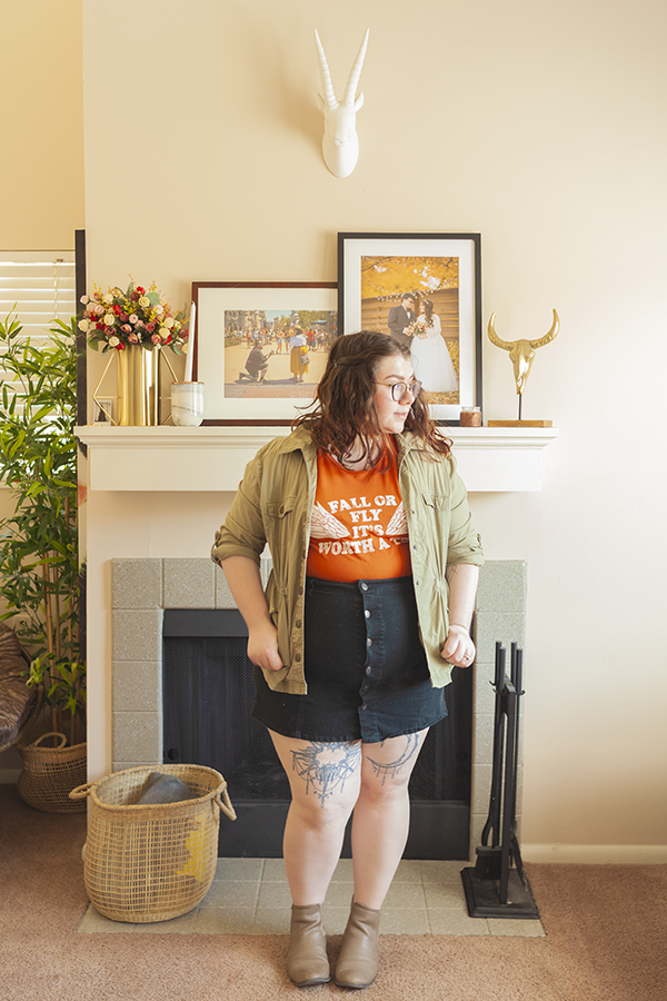 An outfit consisting of an olive green jacket, orange graphic tee with the text "Fall or Fly It's Worth A Try" tucked into a black button down mini skirt and boots.