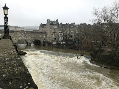 Pultney bridge. The sky above is grey, and large amounts of water are rushing through the arches of the bridge and over the weir.
