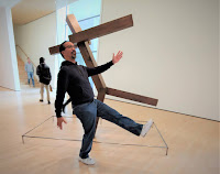 Man walking in an art gallery while trying to align with the action of a wood sculpture of a man walking