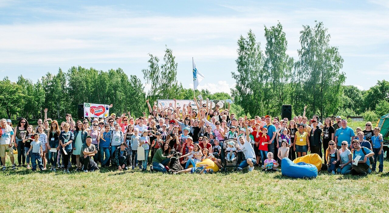A large crowd of Clean Games participants, including adults and children, poses for a group photo outdoors in a grassy field with trees in the background, many smiling and raising their hands in celebration.