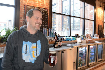 Photo of Kevin Loftis, co-owner of Mother Stewart's Brewing, holding a glass of beer behind the bar.