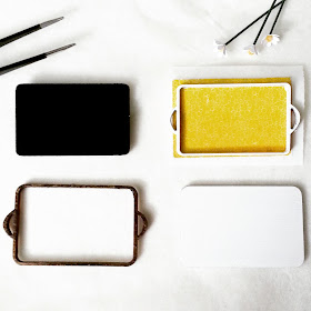 Half-finished one-twelfth scale miniature tray kits: one has a black base and wooden edges, the other a yellow formica base and white edges.