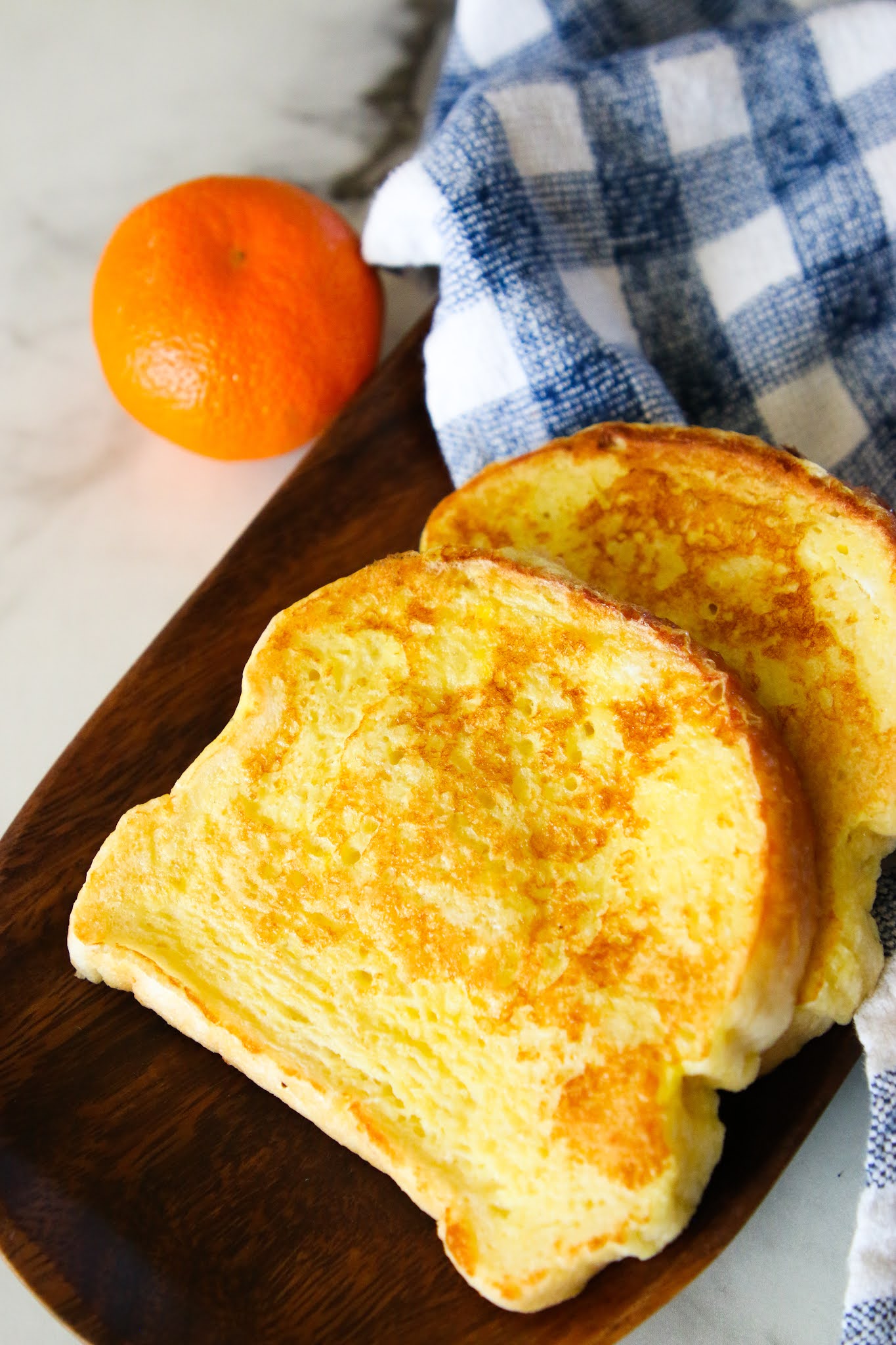 Two pieces of French Toast on a wood plate with a blue dish towel and an orange in the background. There is a for holding a bite of the French Toast.