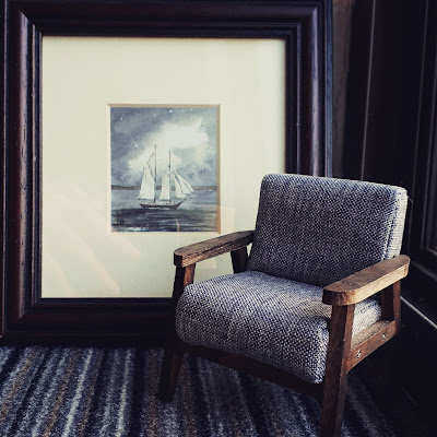 Small framed watercolour of a boat propped up against the wall. In front of it is a piece of carpet tile and a 1/12 scale modern miniature armchair in greys and browns.