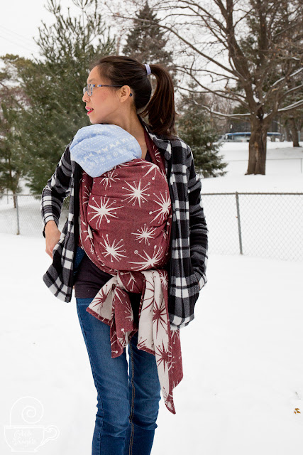 Image of a tan skin bespectacled Asian woman with dark brown hair up in a high pony tail. She's looking over her right shoulder and wearing a sleeping toddler on her front in a maroon and ecru retro star patterned woven wrap carrier underneath a black and white fleece jacket with skinny jeans. They're outside in a winter wonderland with evergreen trees in the background and snow beneath their feet.