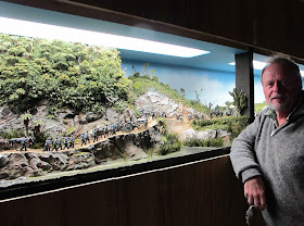 A man poses in front of a diorama of 19th-century soldiers marching in the New Zealand bush.
