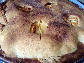 close-up shot of the top of an apple pie. Cinnamon and sugar have carmelized on top of the crust, which is slightly cracked, and apples are visible through some decorative holes on top of the pie