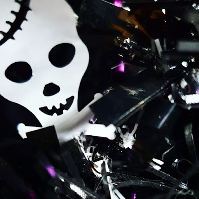 Skull tinsel In the upper left corner is a white foil stylised skull with a crack in its temple, it is attached to black tinsel. The whole thing is ensconced in black synthetic Christmas tree branches, but only the skull is in focus