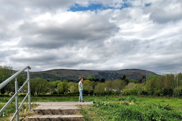 A young blonde haired boy stands up on a raised concrete platform with his hand s in his pockets wearing blue jeans with a grey top. He is staring off to the right hand side surrounded by trees with a mountain off in the distance below a white cloud filled sky with small spots of blue peeping out.