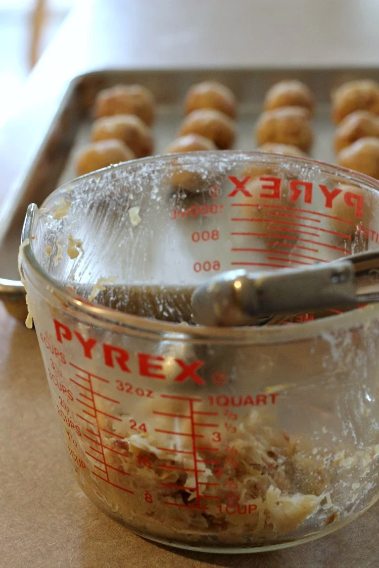 Shaping the cold sauerkraut and ham mixture into balls. Balls on metal baking tray in the foreground and the 4 cup glass measuring cup with a scoop in the front.