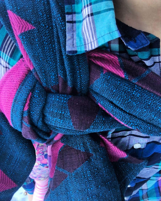 [Image is the close-up of a loop candy cane chest belt finish using a deliciously textured blue and pink woven wrap carrier worn over a jewel-toned plaid shirt. A pink striped sock and confetti print leggings belonging to the child on Mama’s back are peeking out at the bottom.]