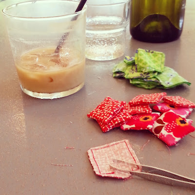 Table in a cafe with an almost-finished glass of iced coffee and a pile of miniature cushions waiting for stuffing. In the foreground is a pair of tweezers.