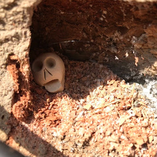 A close up picture of a small, ceramic skull, Skulferatu #49, in the hollow of a crumbling brick below the sign for Nightly Bile Beans.  Photo by Kevin Nosferatu for the Skulferatu Project.