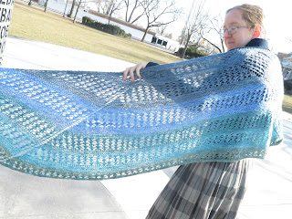 A woman wearing a garter stitch and lace crescent shawl. The shawl is striped in a blue-green gradient. One edge of the shawl is out of frame, with the other end wrapped around her shoulder, with the wingspan held open
