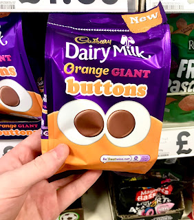 A purple square pouch filled with giant dark brown chocolate buttons with Cadbury in gold font with Dairy Milk in large white font with orange in small orange font and buttons in white font with a photo of two giant circular dark brown chocolate buttons on a white background on a bright background