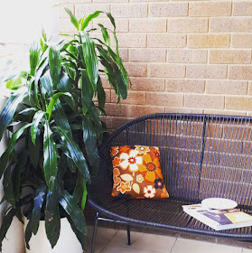 Balcony with two large pot plants next to a mid-century modern wire sofa. On the sofa is a bright flowery cushion, a book and a plate.