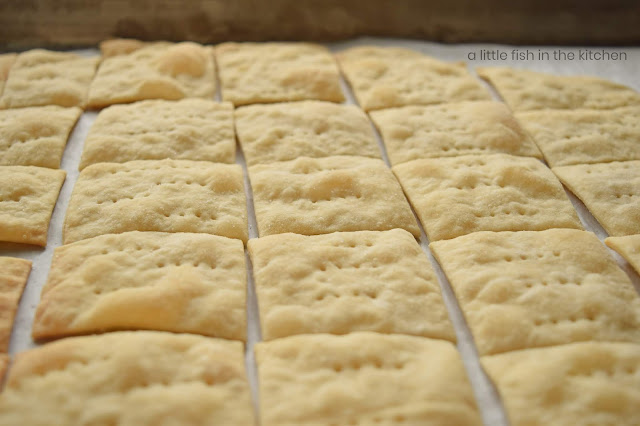 Crisp saltine crackers are cooling on a parchment paper-lined baking sheet. The golden tops of the crackers are slightly puffed and each cracker is pierced with rows of holes from a fork. 