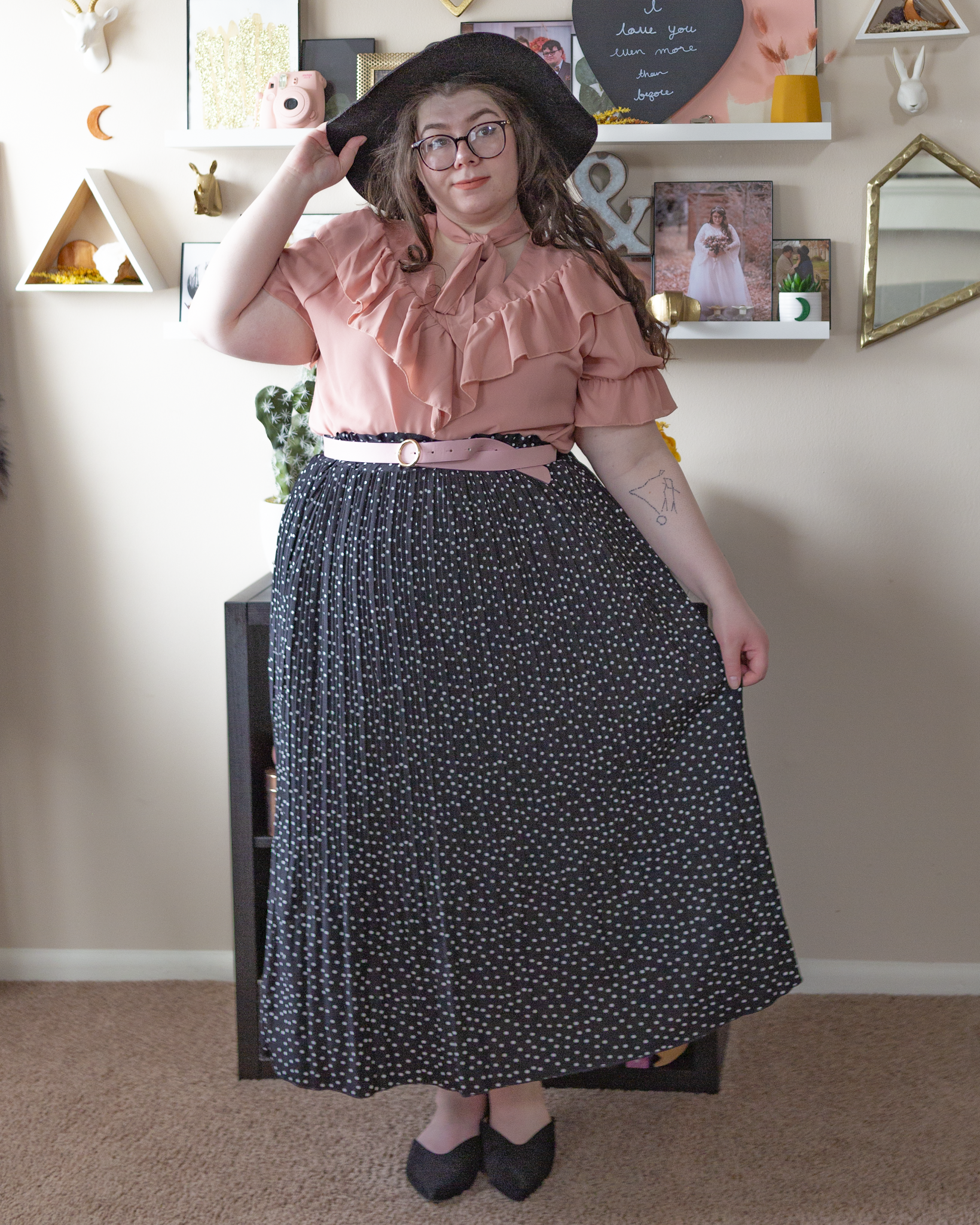 An outfit consisting of a black wide brim fedora, a pastel pink puff sleeve ruffle neckline blouse tucked into a white on black micro swiss dot pleated midi skirt and black pointed toe sling back flats.