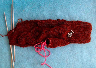 a fingering-weight mitten in progress on double-pointed needles. It has a cable pattern along the back of the hand, and two silver stitch markers clipped in.