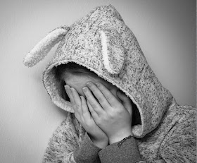 A picture of a child wearing a onesie suit with rabbit ears, with their hands over their eyes, as if they are sad.
