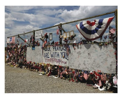 Chain link fence with flowers, toys, flags inserted as tribute to passengers and crew of Flight 93. Two large pieces of plywood painted white have signatures all over them. A large sign in the center says "Our Prayers Are With You."