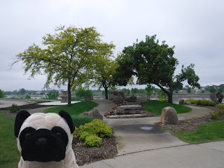 a plush pug appears in front of a small hill with decoratively arranged boulders, the center of which includes a memorial plaque. in the background, the Missouri River is visible