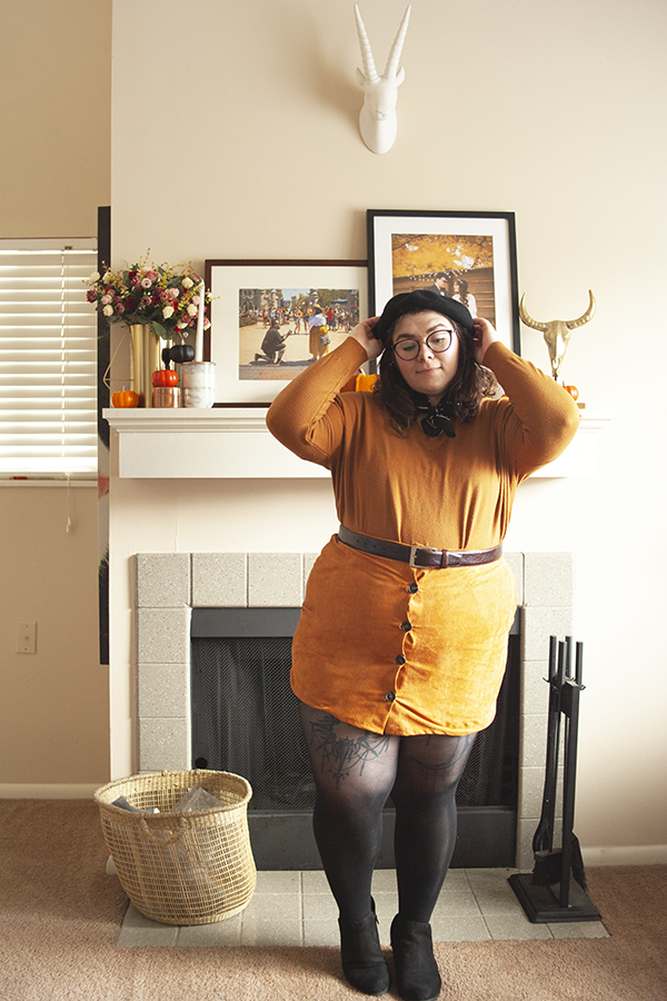 An outfit consisting of a black beret, ochre long sleeve top, brown faux suede button down mini skirt, and black ankle boots.