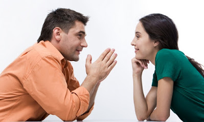 Descrição da imagem #PraCegoVer: Um homem à esquerda, vestido com camisa social, e uma mulher à direita, vestida com blusa de manga curta, ambos sentados, olham nos olhos um do outro. Fim da descrição.