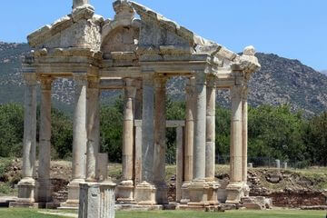Fotografia de ruína de um teatro antigo, onde restam somente as pilastas e uma estrutura do telhado. Construção feita em pedra, numa espécie de praça. Ao fundo se vê vegetação abundante e mais acima, se vê montanhas trespassando o céu, azul claro. Abaixo se pode ver um gramado.