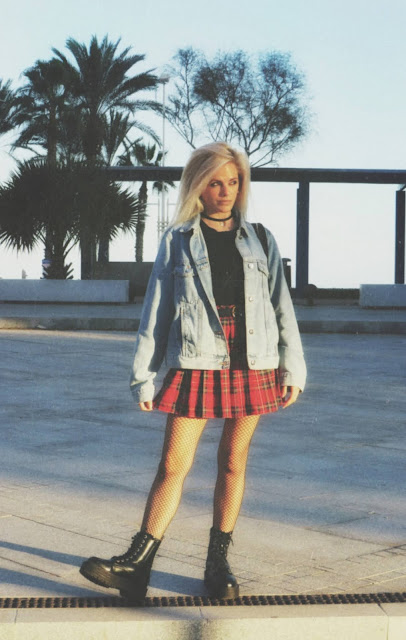 Estilo grunge con camiseta negra, falda de tablas tartán, medias de red grande, botas estilo militar y cazadora vaquera