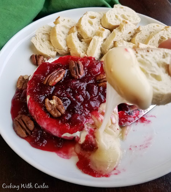 Gooey melty cheese is perfect for toasted bits of baguette. This maple cranberry baked brie is sweet and salty, tarty and gooey. It is perfect for a holiday or just because! plate of baked brie with cranberry sauce and pecans with baguette slice dipped in gooey cheese