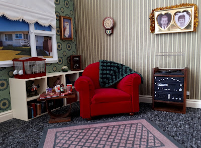 One twelfth scale modern miniature lounge with an overstuffed armchair next to a vintage oak side table and an old-fashioned stereo system. On the wall above the stereo is a picture of a husband and a wife, framed in a double heart-shaped frame, and on the bookcase under the window is an empty bird cage.