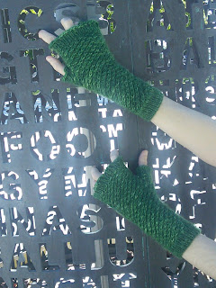  Someone wearing a pair of fingerless mittens. The mittens are knit in a dark green fingering-yarn and use a textured star stitch over the body of the mitten.