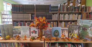 Książki o drzewach i lesie stoją obok siebie na regale. Pośrodku jesienny bukiet kwiatów. Tło: regał z książkami.