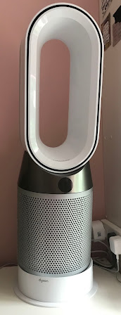 A photograph of a Dyson fan. The top component is a large white hollowed out oval, below this is a dark grey componant with an LED scree in which the top componat sits on. The next component below is a light grey silver cilllinder with lots of holes in it which is where the filters are placed inside. The final component is white with small text with the Dyson logo.