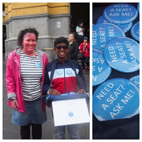 Image: a collage photo. On the left is a woman wearing pink jacket, denim skirt and striped top, red face and curly hair, with an 11 year old boy, wearing red white and navy jacket and glasses. They are smiling, stranding outside Flinders St Station Melbourne. There are a few people in the background. The boy is holding a tray of badges. On the right is a closeup of the badges. They are round and blue with the text 'Need a seat? Ask me!'.