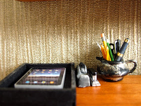 Modern dolls' house scene of a mid-century modern desk with an iPad sitting in an intray, a vintage tape dispenser and stapler and a distressed silver jug holding pens, pencils and scissors.