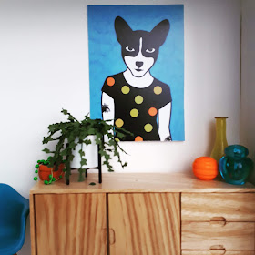 1/12 scale modern miniature scene of a wooden mid-cantury modern sideboard with a string of pearls and an ivy plant on top at one end, and three vases in yellow, orange and teal at the other. Above the sideboard is a print of an animal waering a spotty T shirt in the same colours.