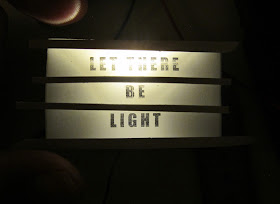 Modern dolls' house miniature light box with the words 'let there be light', half built but lit up, with hand for size reference.