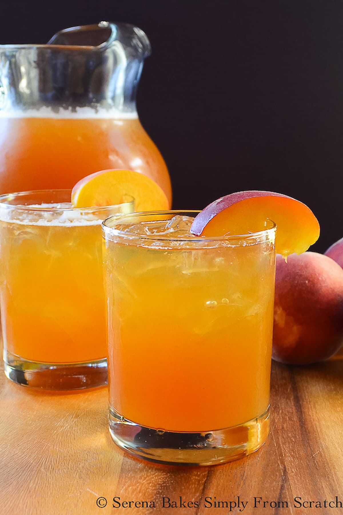 Two glasses of Peach Whiskey Iced Tea with a pitcher in the background on a wooden cutting board.