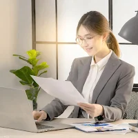 若くて身なりの良いスーツを着た白人女性が、小綺麗なオフィスでノートパソコンと紙の資料を見ながら仕事をしている写真