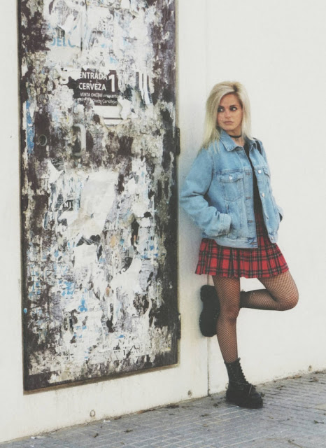 Estilo grunge con camiseta negra, falda de tablas tartán, medias de red grande, botas estilo militar y cazadora vaquera